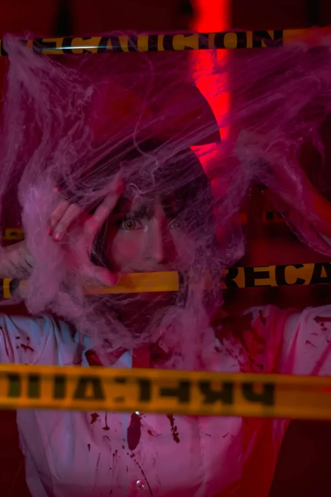 A woman in a creepy costume peers out from behind cobwebs and police tape. The ultimate spooky murder mystery party!